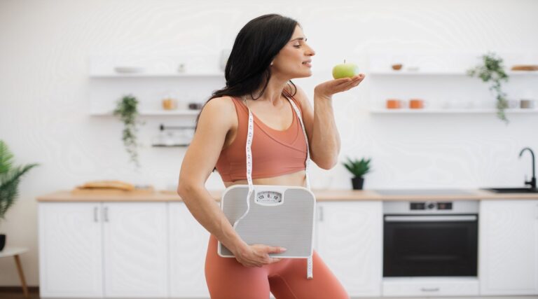Personal trainer posing with apple and weight scales indoors