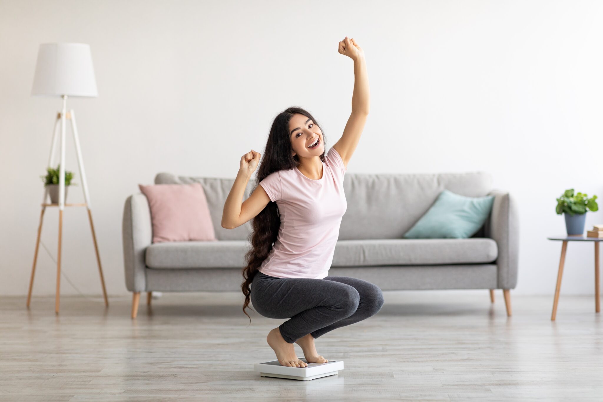 Overjoyed Indian woman sitting on scales, gesturing YES, excited over result of her weight loss diet at home