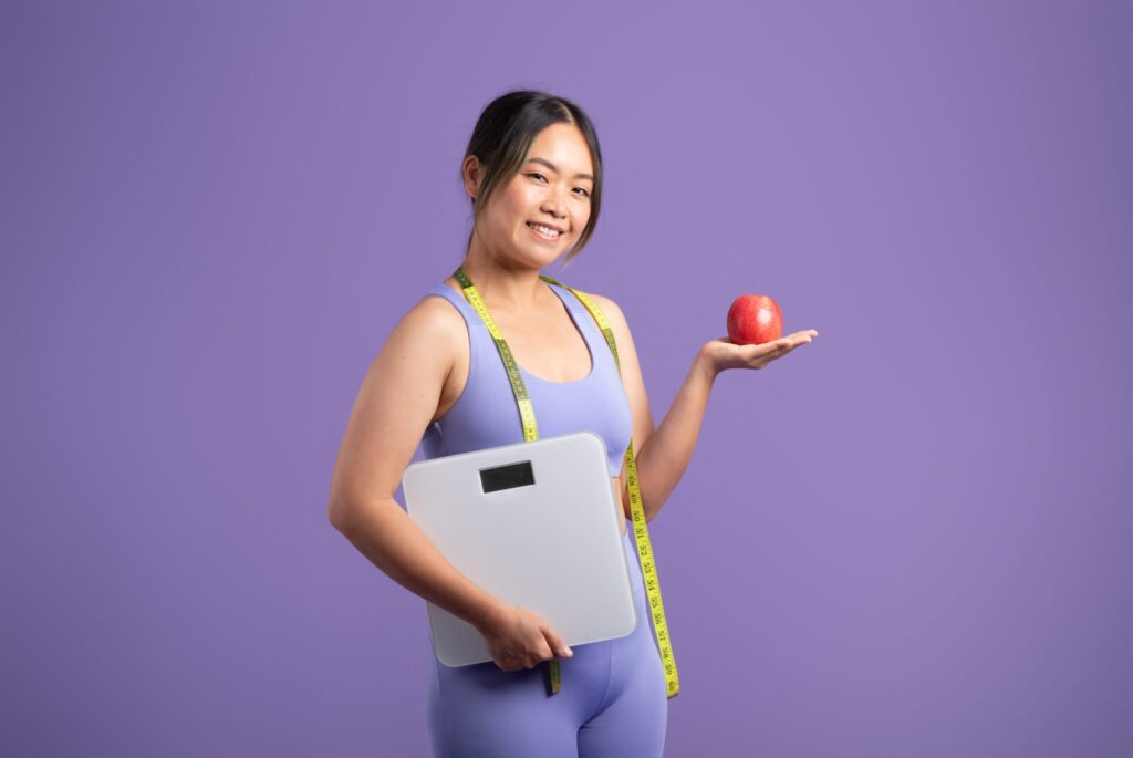 Smiling asian woman holding scales and apple, purple studio background