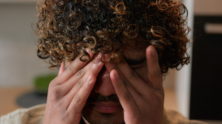 Close up of an ill and tired man with curly hair rubbing dry eyes