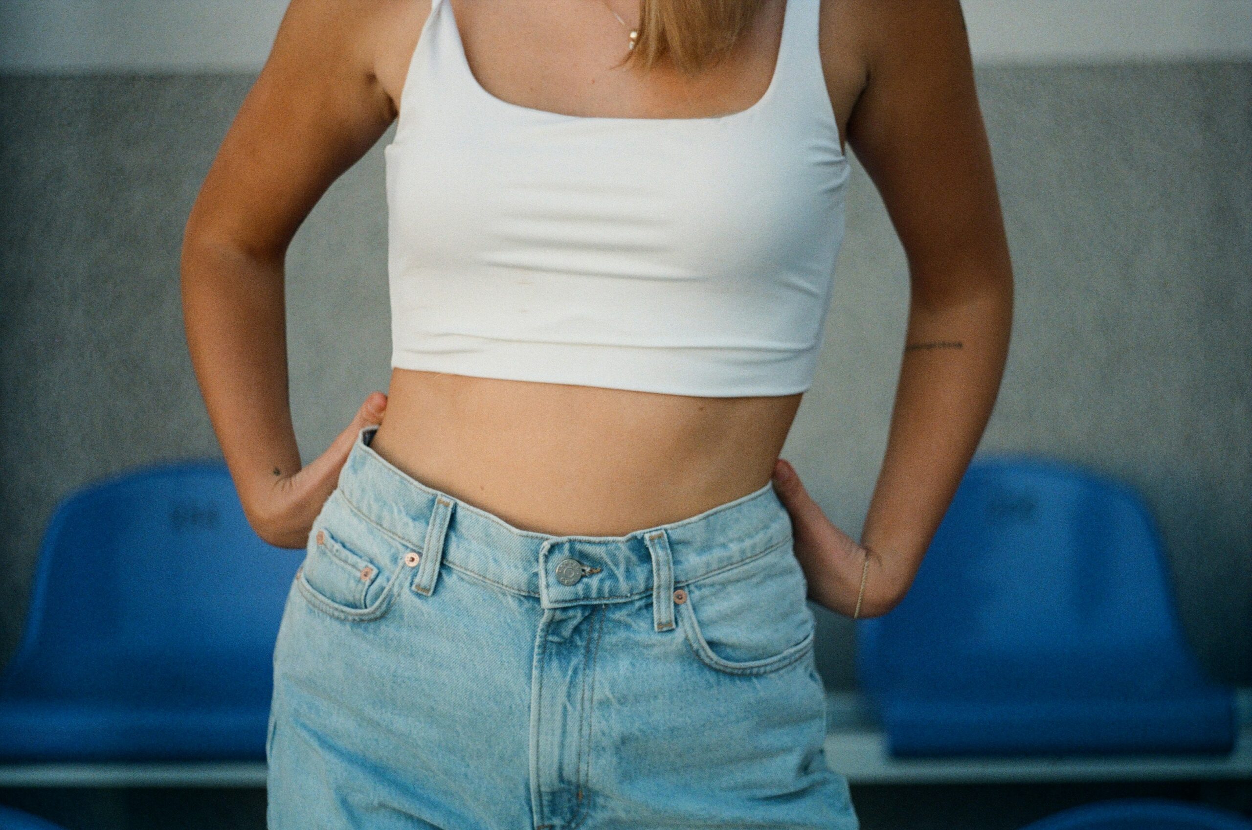 Person in white crop top and jeans standing in front of blue plastic chairs.